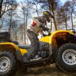 Horizontal motion portrait of a man in gray sport jacket and safety helmet and goggles driving mud-covered yellow ATV 4x4 quad bike with dirt spinning of the wheels.