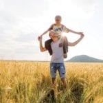 A young father with his little son walking in the wheat field at sunset in a warm summer day