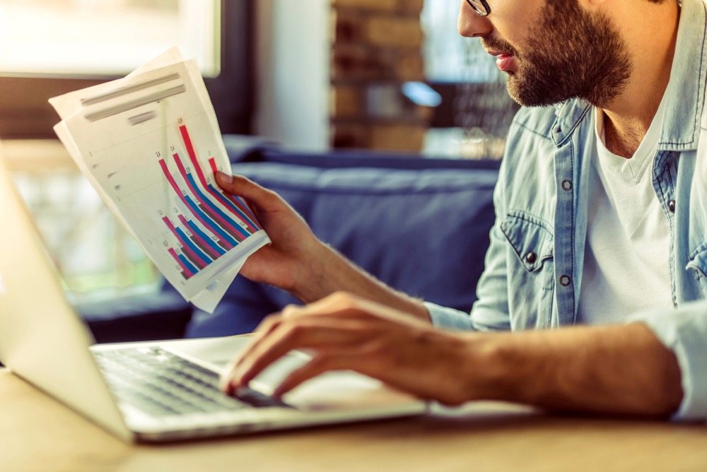 Cropped image of handsome businessman in casual wear using a laptop and examining documents while working in the office