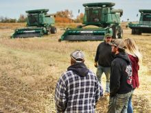 Group of people standing outside on farm field in Canada with agricultural machinery in background during harvest season. Pic: AJ_Watt/E+/Getty Images