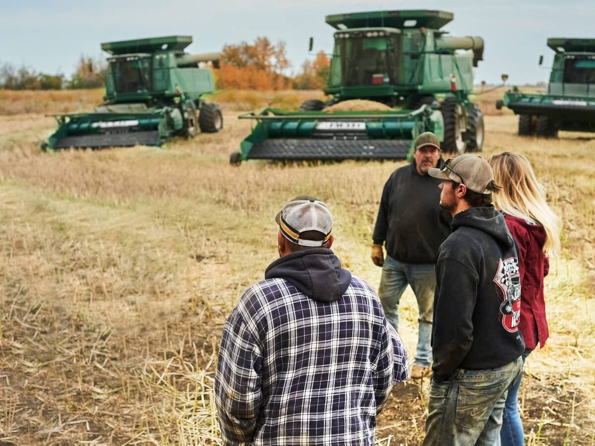 Group of people standing outside on farm field in Canada with agricultural machinery in background during harvest season. Pic: AJ_Watt/E+/Getty Images