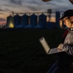 A focused male farmer sits in a car trunk, working on his laptop against a twilight sky, with silos and a lush field in the background. Pic: SimonSkafar/E+/Getty Images