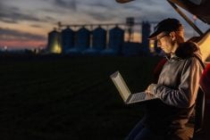 A focused male farmer sits in a car trunk, working on his laptop against a twilight sky, with silos and a lush field in the background. Pic: SimonSkafar/E+/Getty Images