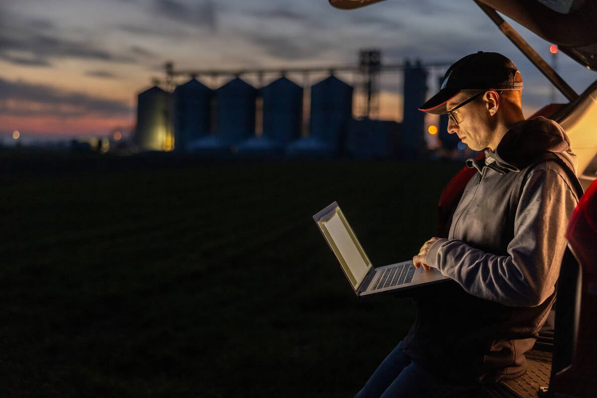 A focused male farmer sits in a car trunk, working on his laptop against a twilight sky, with silos and a lush field in the background. Pic: SimonSkafar/E+/Getty Images