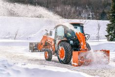 A new orange utility tractor (Kubota L70) with a snowblower attachment at work on a rural lane